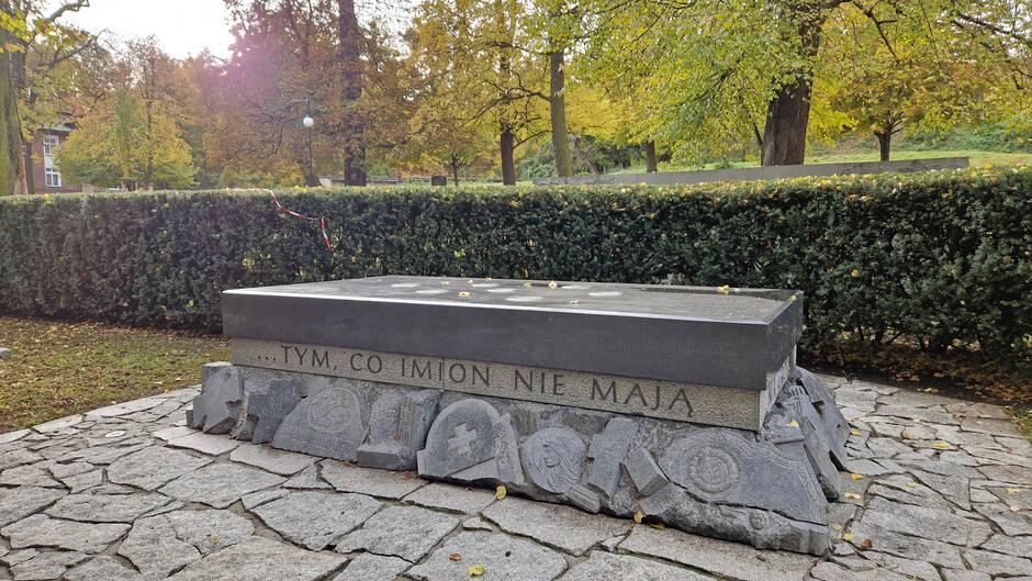 Płaska podwyższona płyta kamienna. U dołu stylizowane fragmenty połamanych nagrobków