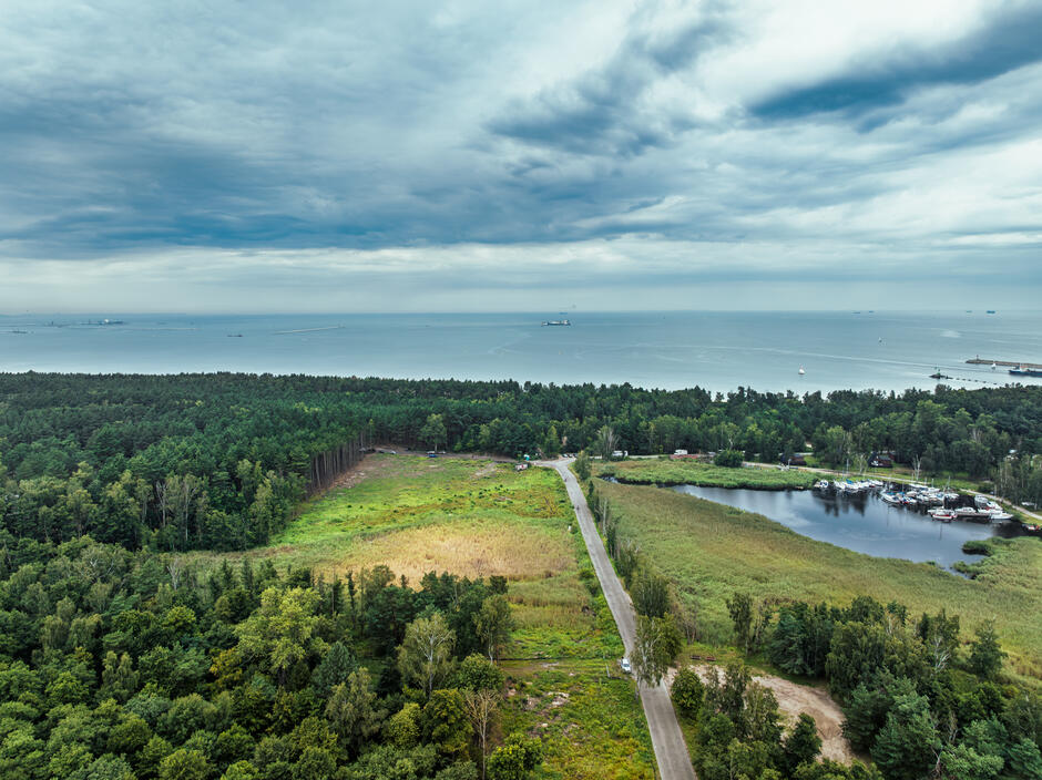 Panorama nadmorskiego krajobrazu ukazuje zalesiony pas terenu oddzielający morze od lądu, z widocznym fragmentem jeziora i mariną. Na horyzoncie widać statki na spokojnej wodzie, a niebo zasnute chmurami podkreśla chłodny klimat wybrzeża.
