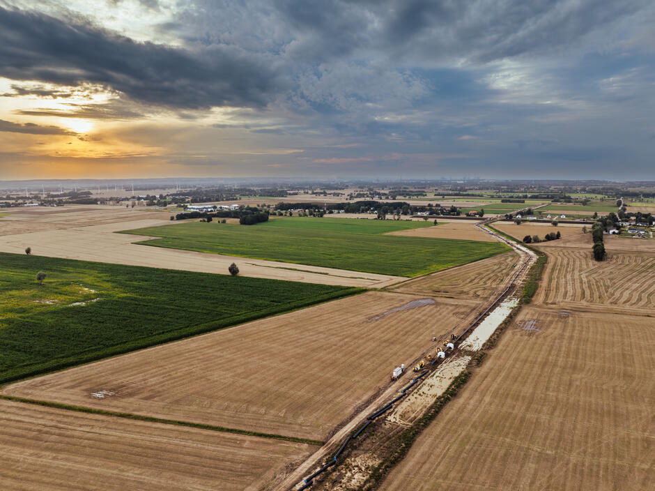 Szeroki krajobraz rolniczy rozciąga się pod zachmurzonym niebem, a przez pola biegnie linia budowy nowej infrastruktury. Wzdłuż wykopu widoczne są maszyny i rurociągi, które tworzą kontrast z naturalnymi barwami pól i zachodzącym słońcem.
