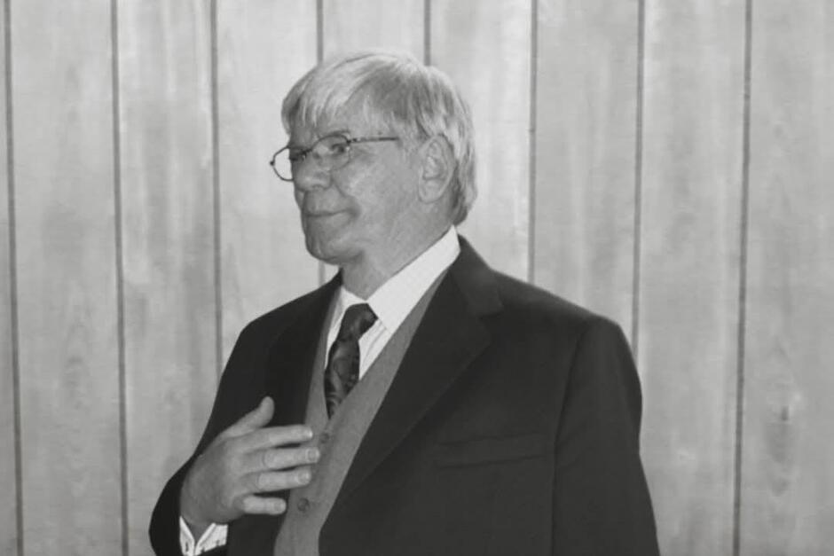 Na zdjęciu widać starszego mężczyznę w garniturze, kamizelce i krawacie, stojącego na tle drewnianej ściany. Ma siwe włosy, okulary i spokojny wyraz twarzy, a jego dłoń spoczywa na piersi w geście zamyślenia lub skupienia.