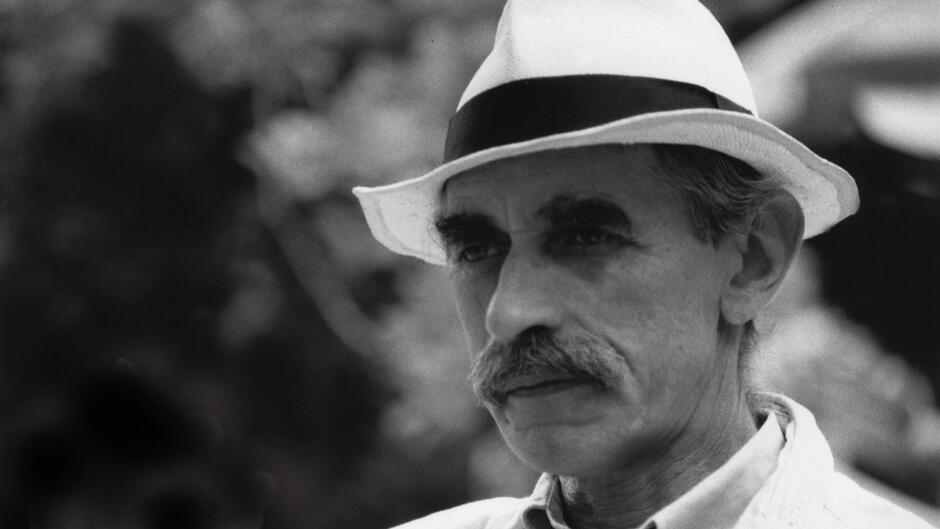 Mężczyzna o szczupłej twarzy, w jasnym kapeluszu, z wąsem.