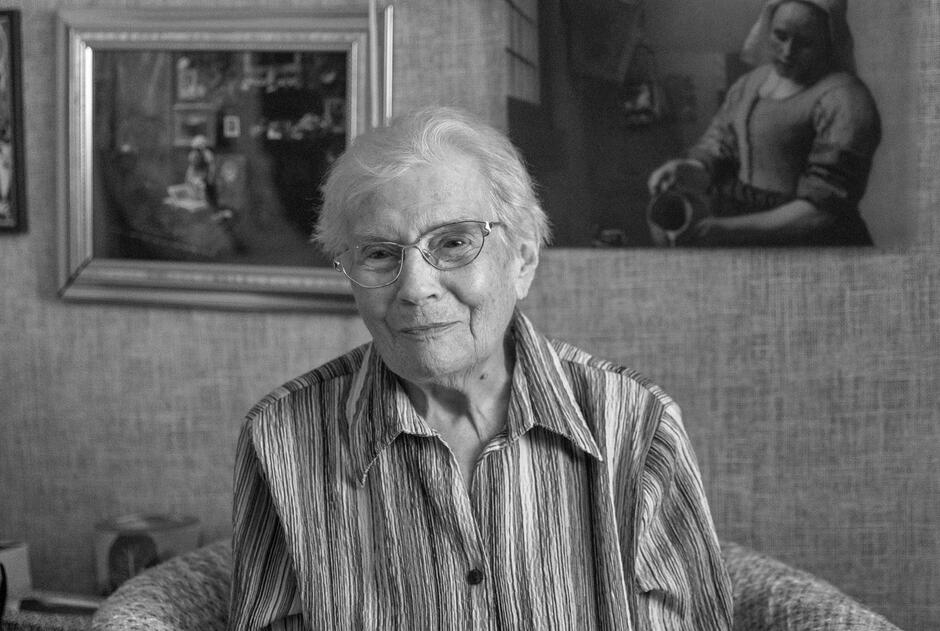Na zdjęciu widać uśmiechniętą starszą kobietę z krótkimi siwymi włosami i okularami, siedzącą w fotelu. Ubrana jest w koszulę w paski, a za nią wiszą obrazy w ramach, co nadaje wnętrzu spokojny, domowy charakter.