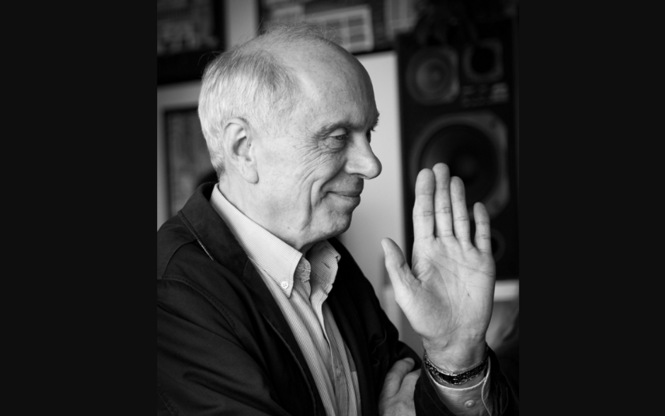 Na czarno-białym zdjęciu widzimy starszego mężczyznę w półprofilu, ubranego elegancko w koszulę i marynarkę, który z uśmiechem unosi rękę w geście powitania lub pożegnania. W tle widać głośniki i rozmyte elementy wystroju wnętrza