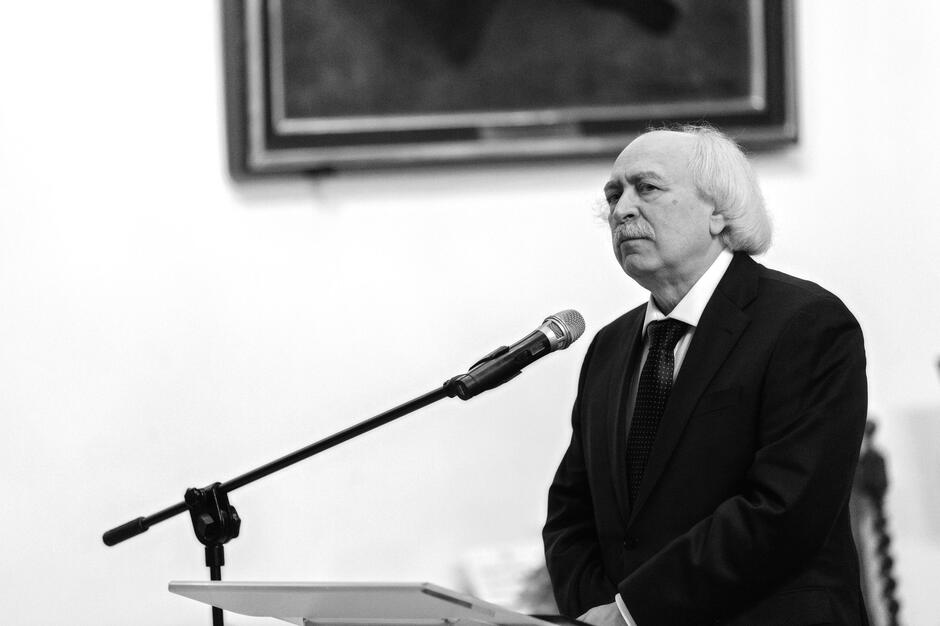 Na czarno-białym zdjęciu widzimy elegancko ubranego mężczyznę w średnim wieku, przemawiającego do mikrofonu podczas uroczystości – jego poważna mina i postawa sugerują, że jest to ważne wystąpienie. Tło i ustawienie wskazują na akademickie lub kulturalne wydarzenie, być może związane z wręczeniem nagrody lub laudacją.