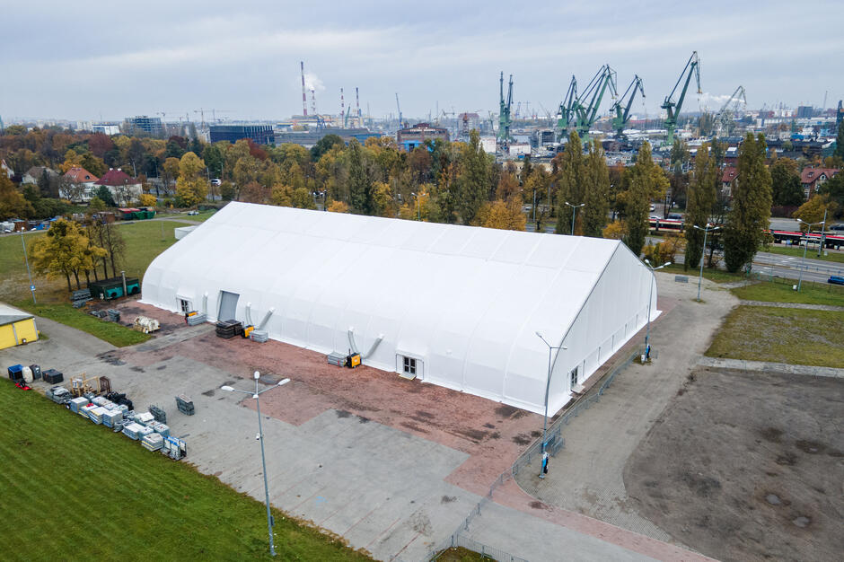 biała wielka hala na betonowym placu, wzdłuż stoją pojazdy techniczne i samochody