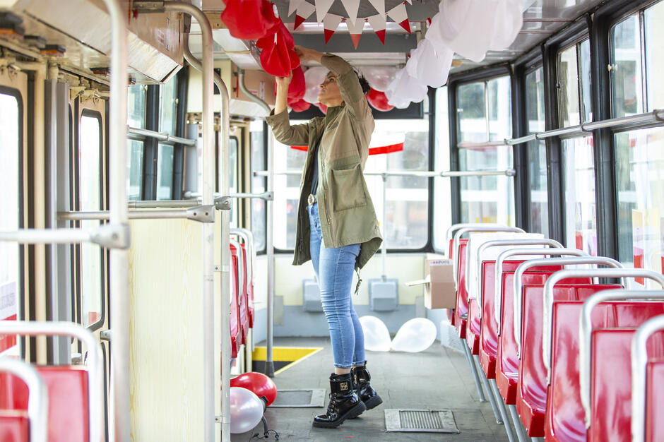 Kobieta w zielonej kurtce i dżinsach wiesza czerwone i białe ozdoby wewnątrz tramwaju. Wnętrze pojazdu wypełniają balony i chorągiewki w barwach narodowych, przygotowywane z okazji święta niepodległości.
