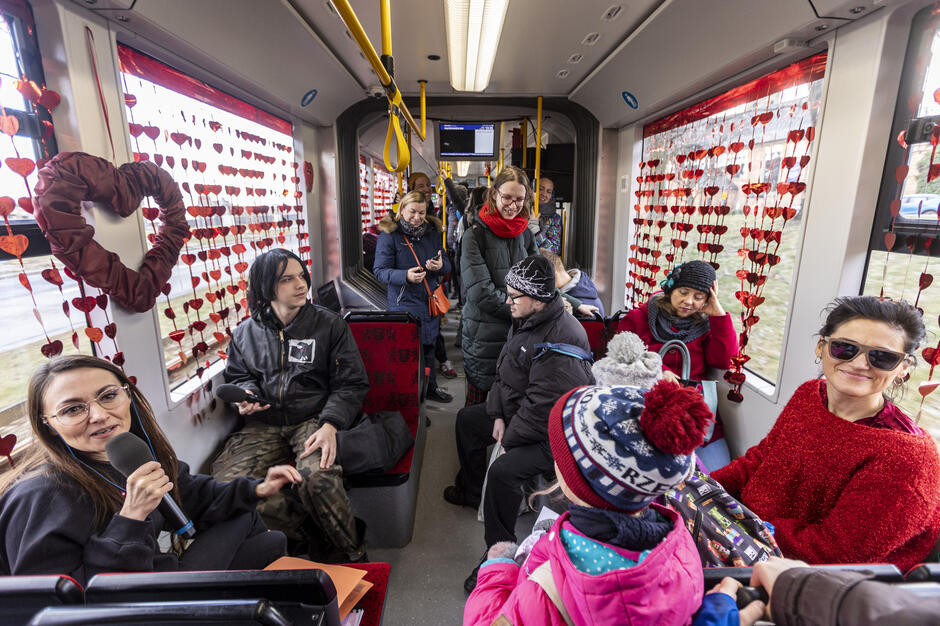 Grupa pasażerów jedzie tramwajem udekorowanym czerwonymi sercami i wstążkami z okazji Walentynek. Wnętrze pojazdu wypełniają rozmowy, uśmiechy i ciepła atmosfera wspólnego świętowania.
