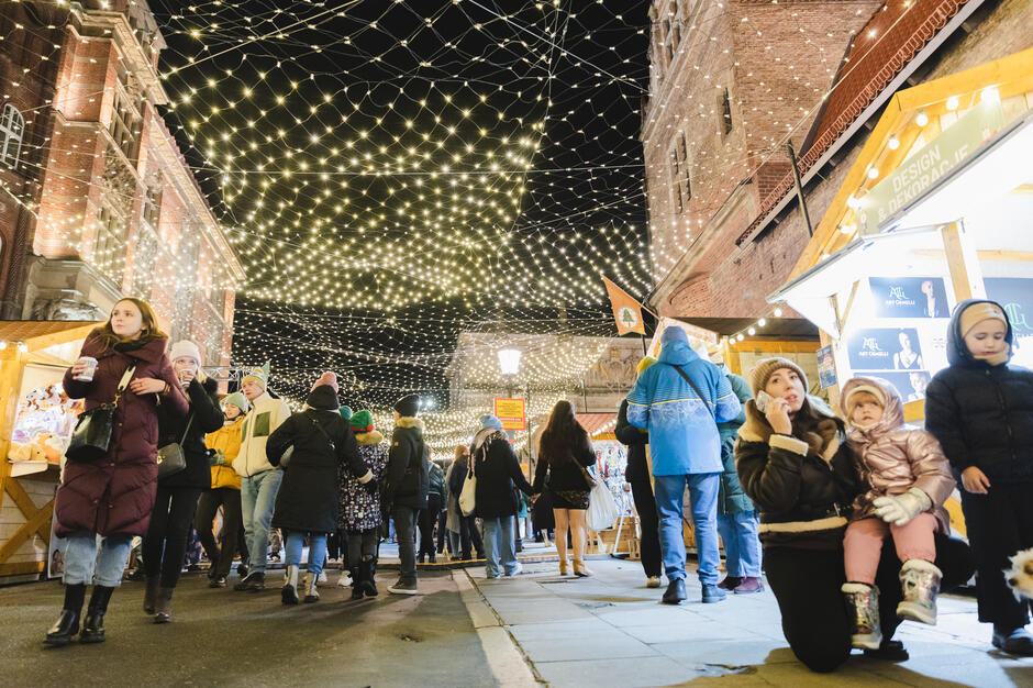 Tłum spaceruje wśród świątecznych stoisk rozświetlonych gęstą siecią lampek, tworzących nad ulicą złocisty baldachim. W powietrzu unosi się atmosfera zimowego jarmarku – ludzie z kubkami gorących napojów podziwiają ozdoby i cieszą się wspólnym czasem. 