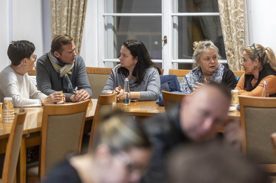 Grupa mieszkańców siedzi przy stole w sali spotkań, prowadząc rozmowę na temat organizacji wydarzeń w mieście. Uczestnicy dyskusji wymieniają opinie w spokojnej atmosferze, mając przed sobą napoje i notatki.
