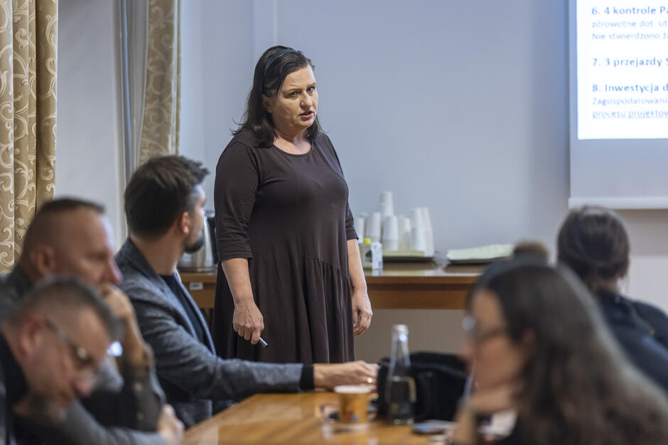 Kobieta prowadzi prezentację podczas spotkania, omawiając punkty wyświetlone na ekranie. Uczestnicy siedzący przy stole słuchają jej wystąpienia, notując i obserwując slajdy.
