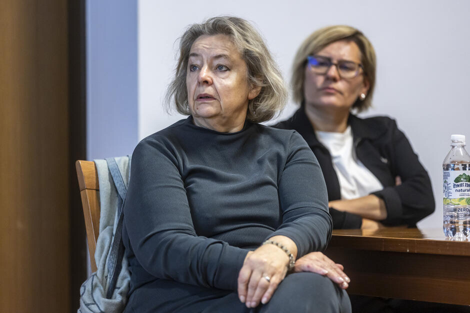 Dwie kobiety uczestniczą w spotkaniu, uważnie słuchając prezentowanych informacji. Siedzą przy stole w sali konferencyjnej, zachowując skupienie i powagę. 