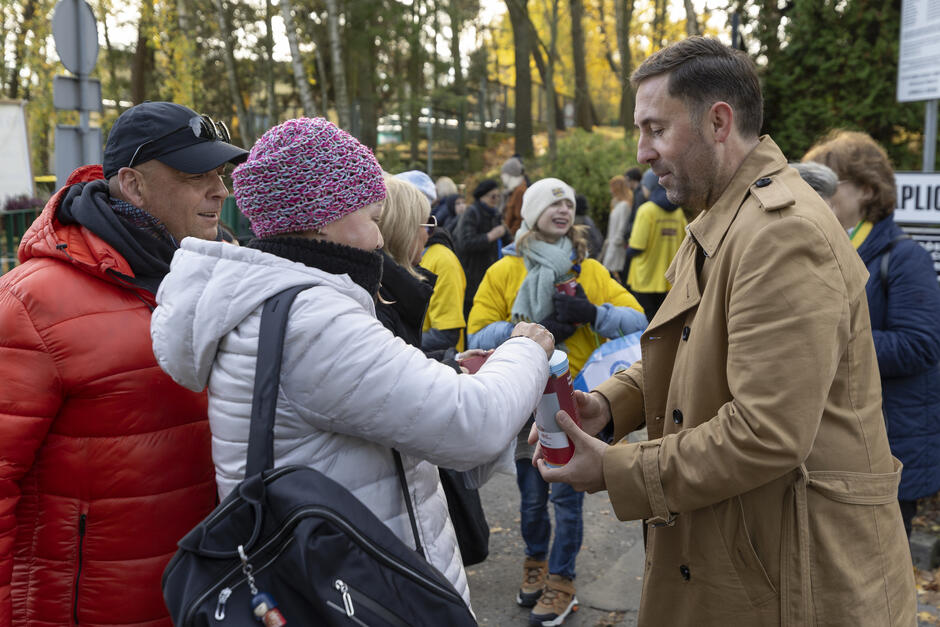 Mężczyzna w jasnym prochowcu zbiera wśród przechodniów pieniądze do puszki