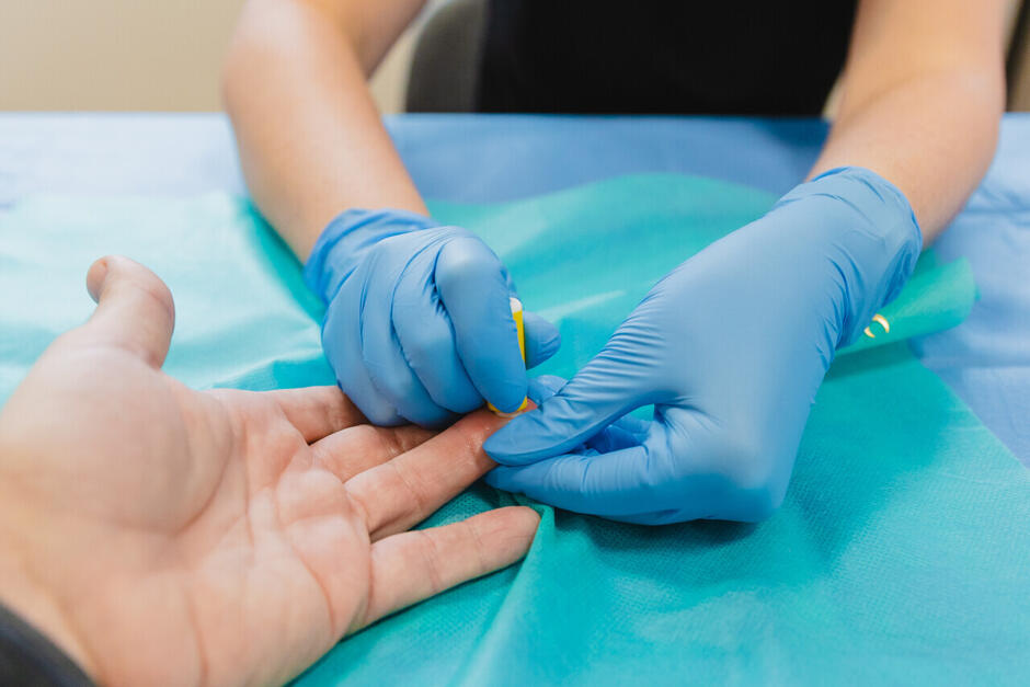 Zdjęcie przedstawia pracownika medycznego wykonującego nakłucie palca na ręce pacjenta w celu pobrania próbki krwi.
