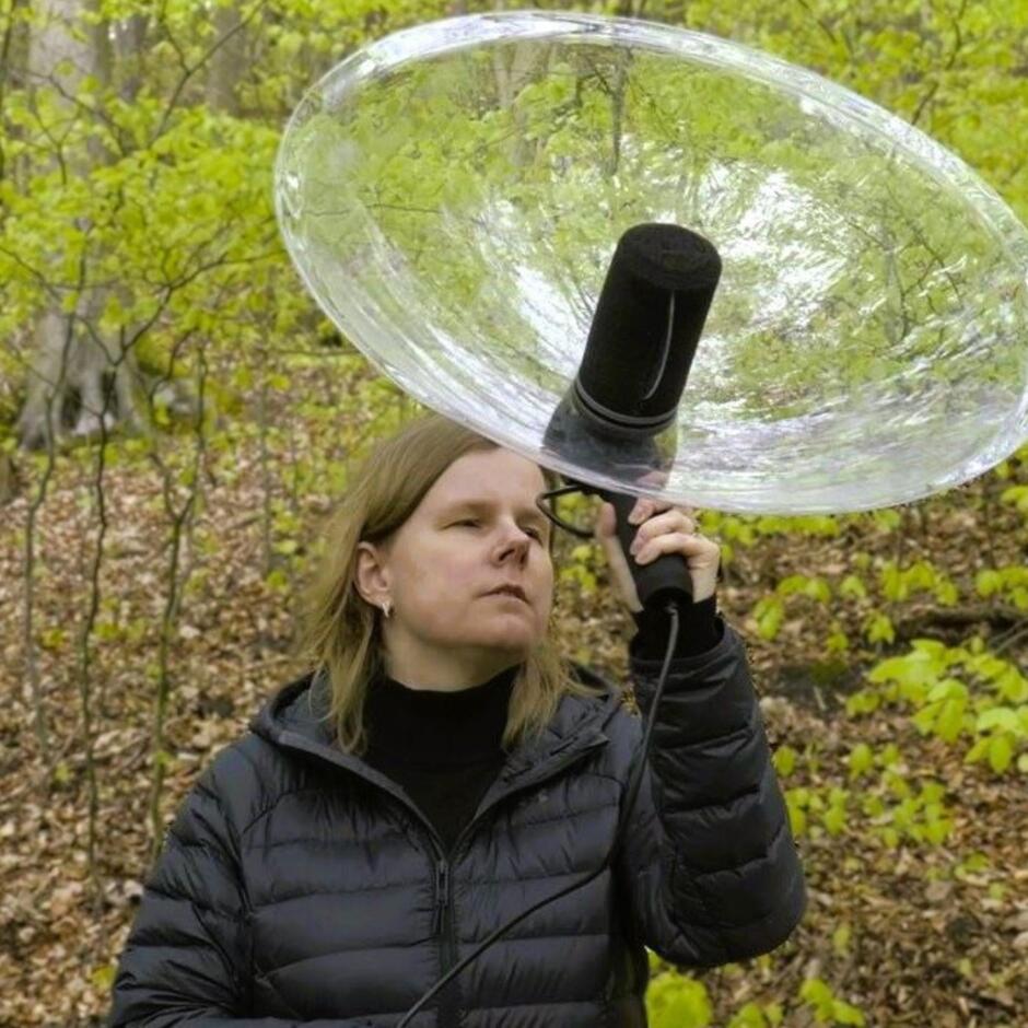 Kobieta w czarnej kurtce trzyma w lesie duży mikrofon z przezroczystą paraboliczną osłoną, nasłuchując dźwięków otoczenia. W tle widać drzewa z młodymi, jasnozielonymi liśćmi i warstwę suchych liści na ziemi.
