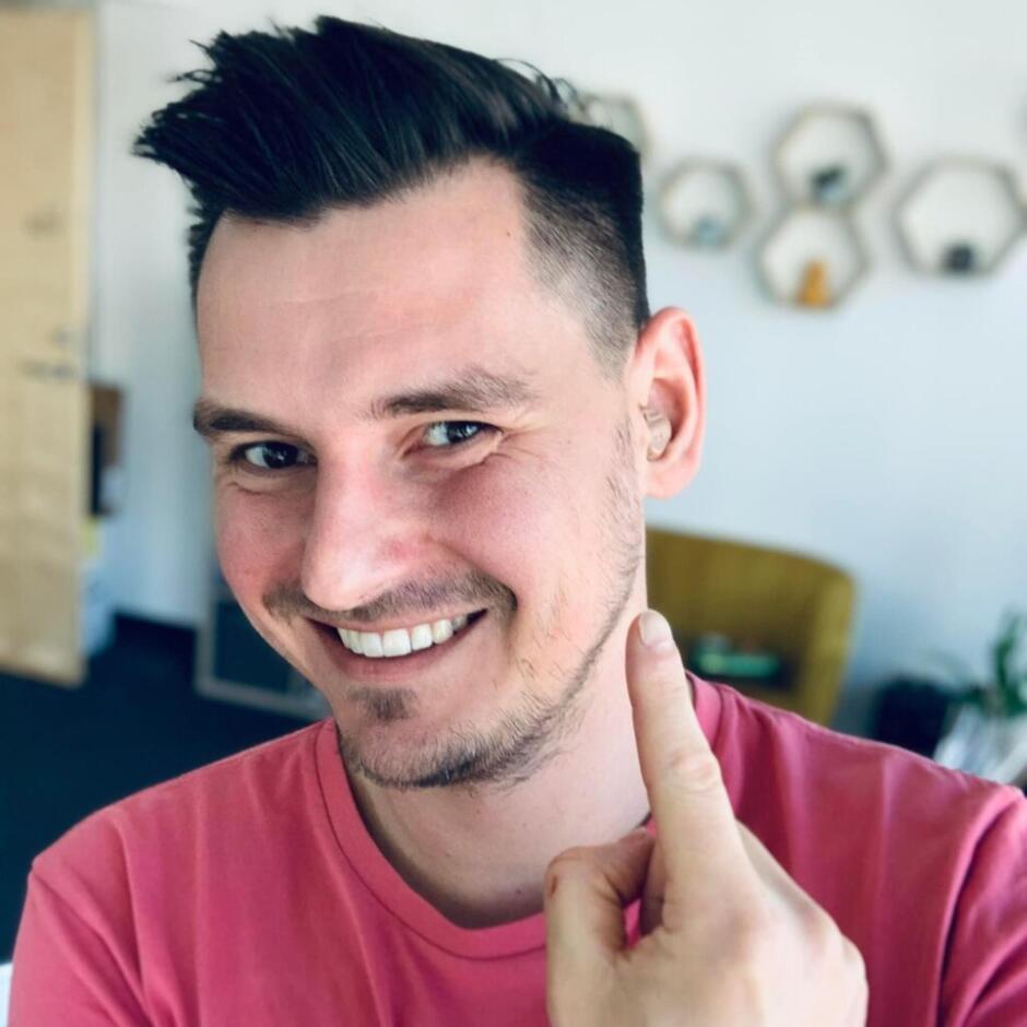 Na zdjęciu uśmiechnięty mężczyzna w różowej koszulce wskazuje palcem na swoje ucho, w którym ma dyskretne urządzenie słuchowe. W tle widać jasne wnętrze z geometrycznymi półkami na ścianie i żółtym fotelem.
