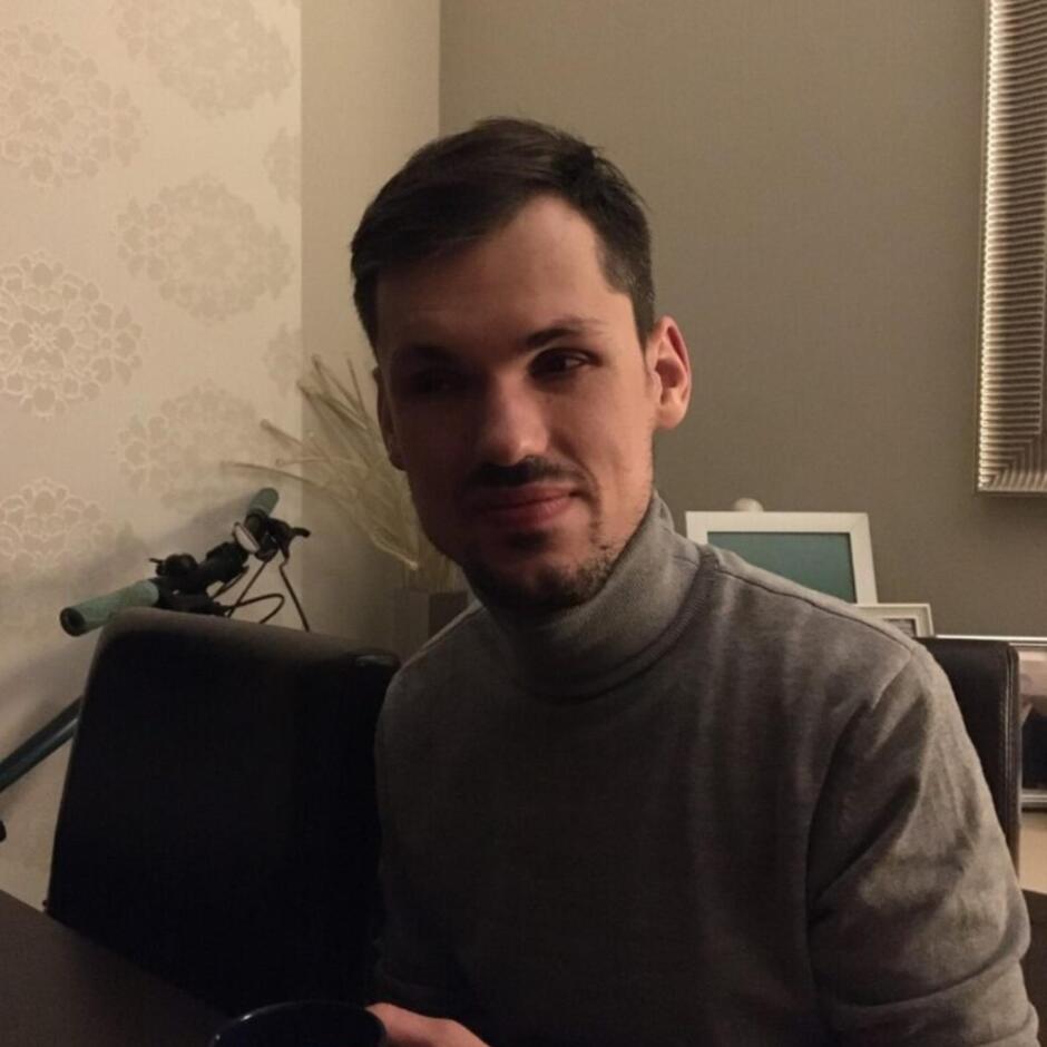 Mężczyzna w szarym golfie siedzi przy stole, trzymając w dłoni kubek. W tle widać rower, ramkę na zdjęcie i jasną ścianę z delikatnym wzorem.
