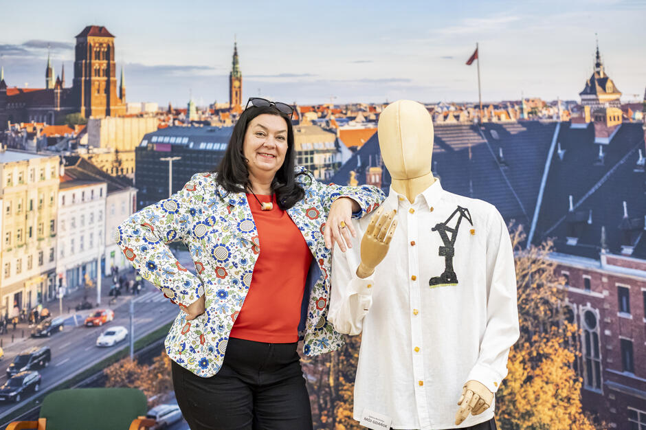 Na zdjęciu widać kobietę, która uśmiecha się, stojąc obok manekina, który ma na sobie koszulę ozdobioną unikalnym wzorem przedstawiającym dźwig. W tle rozciąga się panorama Gdańska z charakterystyczną architekturą, w tym wieżą kościoła i dachami budynków.
