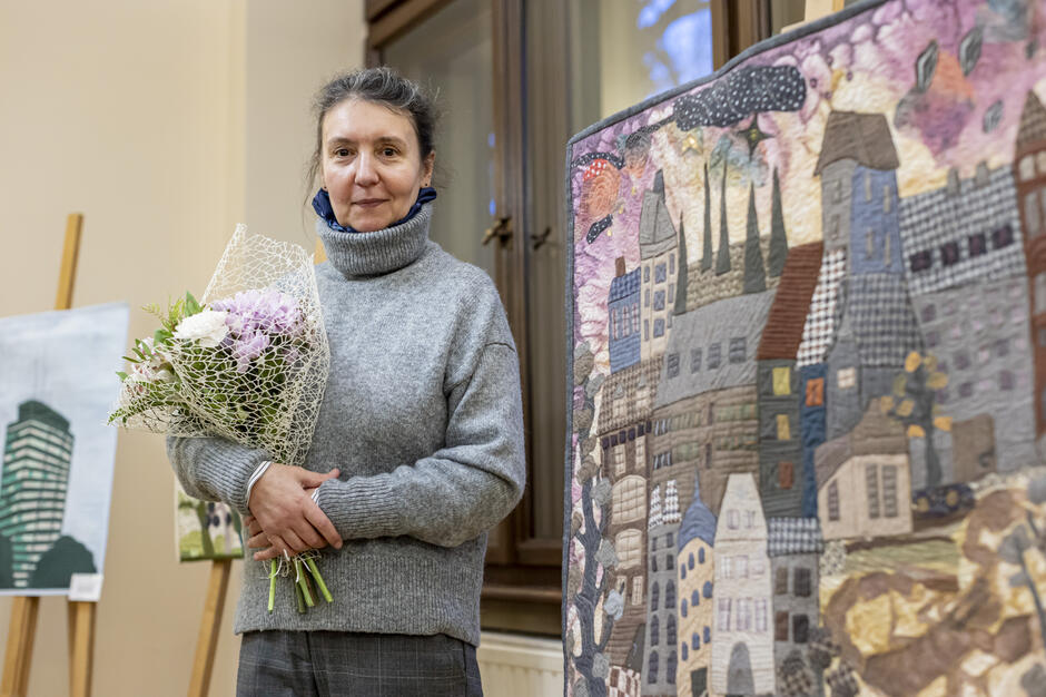 Na zdjęciu widzimy kobietę trzymającą bukiet kwiatów, ubrana w szary sweter i szare spodnie, stojącą obok patchworkowego obrazu przedstawiającego wizerunek miasta. Tło to wnętrze sali, w której wystawione są inne prace artystyczne.
