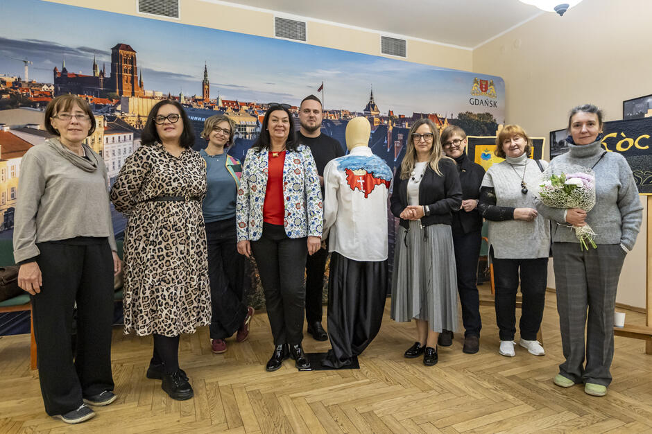 Na zdjęciu grupa osób stoi przed manekinem, który ma na sobie koszulę z haftowanym wzorem przedstawiającym mapę Polski. W tle widać panoramę Gdańska, a uczestnicy wystawy są ubrani w różne stylizacje, prezentując różnorodność uczestników wydarzenia.
