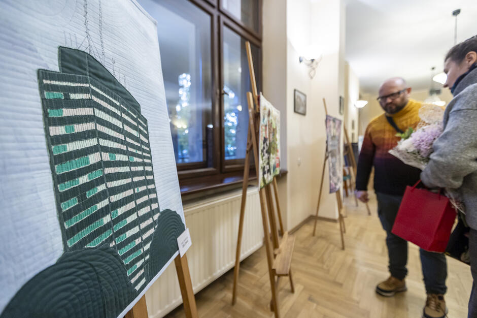 Na zdjęciu widać uczestników wystawy patchworków, którzy oglądają jedno z dzieł przedstawiających nowoczesny budynek. W tle stoją kolejne prace artystyczne, a atmosfera jest pełna twórczego zaangażowania, z wyraźnym akcentem na detale w tekstylnych obrazach.
