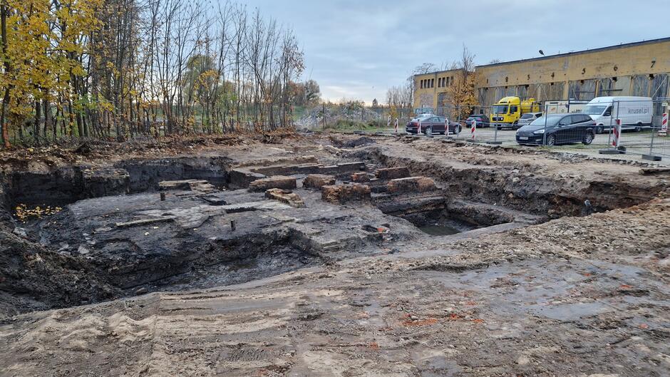 Ceglane fundamenty w ciemnej ziemi, z tyłu żółty budynek dawnej zajezdni i parkujące przed nim samochody