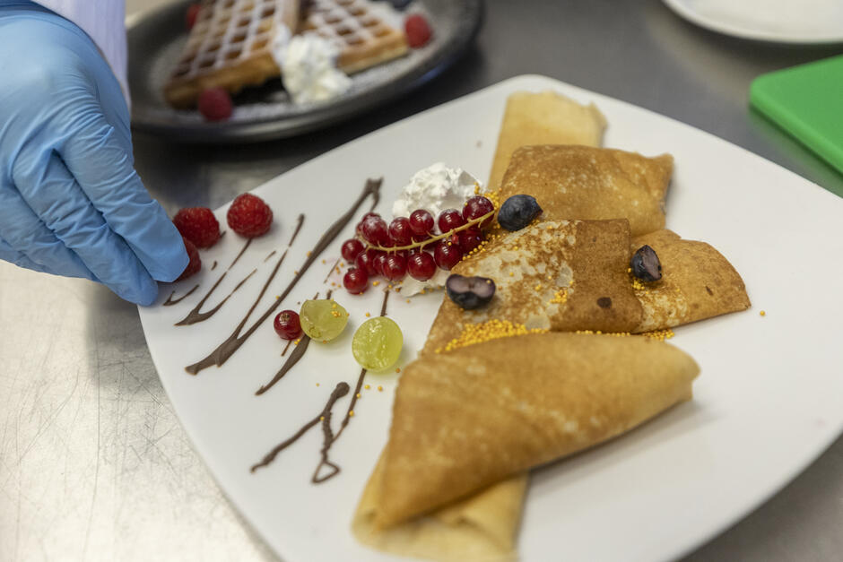 Na talerzu znajduje się deser z naleśnikami udekorowanymi owocami — malinami, porzeczkami, winogronami i borówkami — oraz czekoladowym sosem i bitą śmietaną. W tle widać gofry, a osoba w rękawiczce układa składniki, dbając o estetykę podania.
