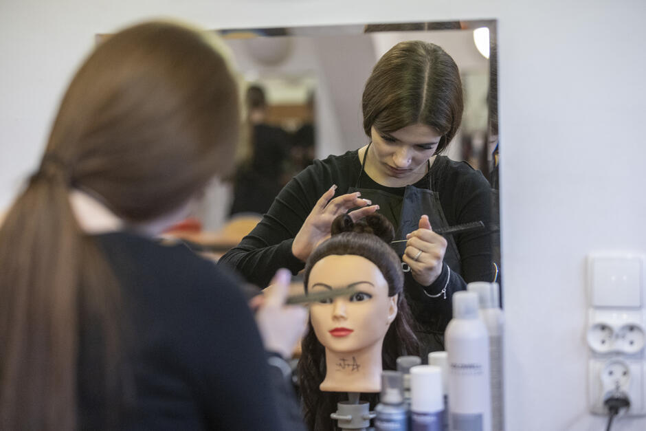 Uczennica wykonuje fryzurę na głowie treningowej, skupiając się na precyzyjnym upięciu włosów. W lustrze odbija się jej sylwetka oraz stanowisko z kosmetykami i akcesoriami fryzjerskimi.
