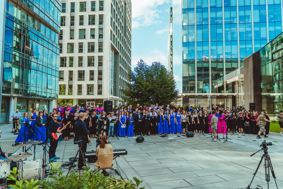 Około 150 osób stoi na dziedzińcu między nowoczesnymi budynkami i śpiewa.