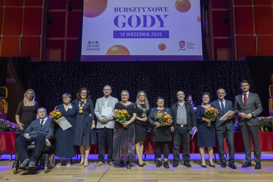 Kilkanaście osób pozuje do zdjęcia na scenie, nad nimi na ekranie napis Bursztynowe gody . Jeden z mężczyzn siedzi na wózku inwalidzkim.