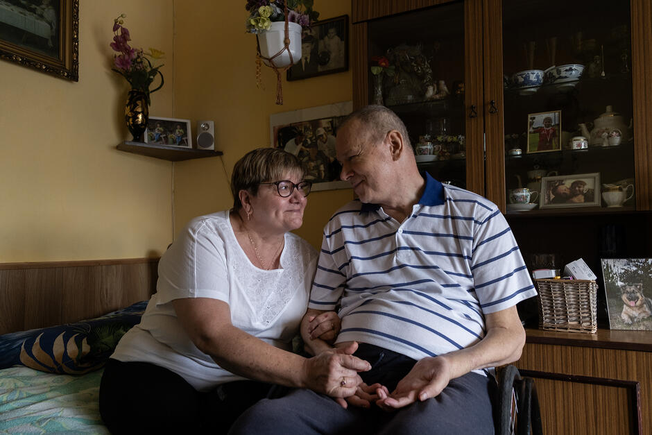 Kobieta siedzi na łóżku, obok mężczyzna siedzi na wózku inwalidzkim. Trzymają się za ręce i patrzą na siebie.