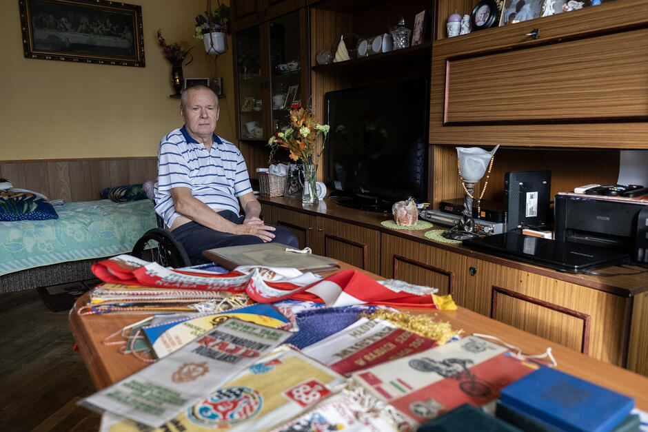 Mężczyzna siedzi w pokoju na wózku inwalidzkim i patrzy na stolik, na którym leżą proporczyki i wycinki z gazet.