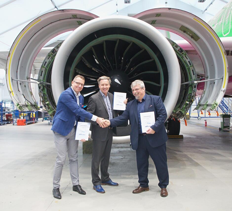 Trzej mężczyźni potwierdzają umowę uściskiem dłoni. W tle jest wielki silnik samolotu pasażerskiego, widoczny od strony wirnika 