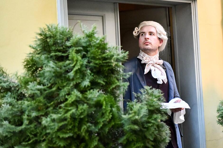Mężczyzna w historycznym stroju z peruką i koronkowym żabotem stoi w drzwiach budynku, spoglądając w górę. Scena przypomina rekonstrukcję z epoki, a jego elegancki ubiór i poważny wyraz twarzy podkreślają teatralny charakter przedstawienia.
