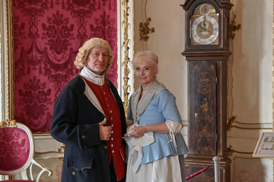 Para w strojach z XVIII wieku pozuje w eleganckim wnętrzu z ozdobnymi tapetami i zabytkowym zegarem. Ich ubiór i otoczenie tworzą atmosferę dawnego dworu, podkreślając historyczny charakter wydarzenia.
