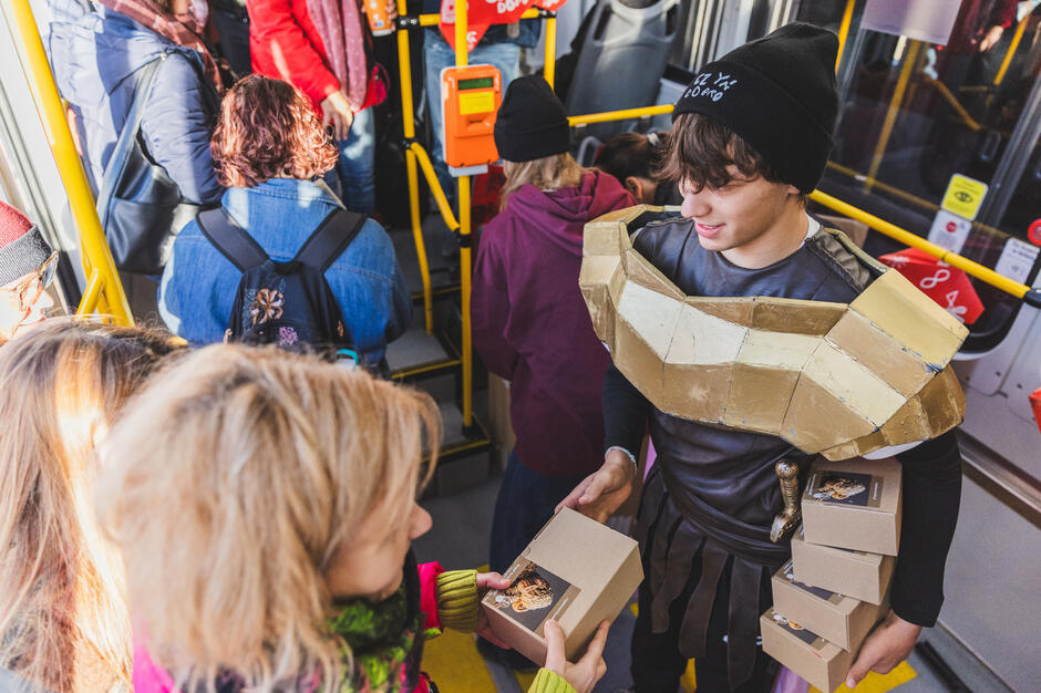Młody mężczyzna przebrany za rzymskiego żołnierza rozdaje w tramwaju pudełka z rogalami.