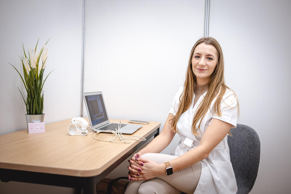 Kobieta w białym uniformie medycznym siedzi przy biurku z laptopem, na którym widoczny jest wykres diagnostyczny. Na stole leżą także urządzenie diagnostyczne, smartfon i doniczka z rośliną. Kobieta uśmiecha się do kamery, siedząc w jasnym, minimalistycznym pomieszczeniu.