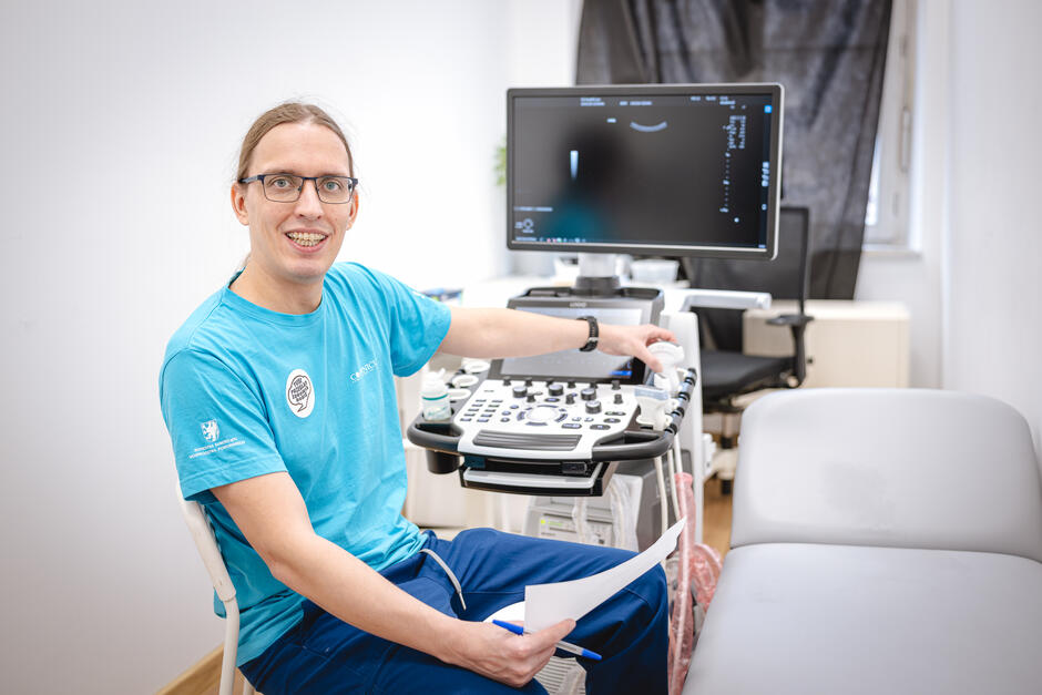 Mężczyzna w okularach i niebieskiej koszulce siedzi w gabinecie medycznym obok aparatu USG. Uśmiecha się do aparatu, trzyma w ręku kartkę i długopis, a drugą ręką obsługuje urządzenie diagnostyczne. W tle widać monitor z obrazem z USG oraz leżankę dla pacjentów.