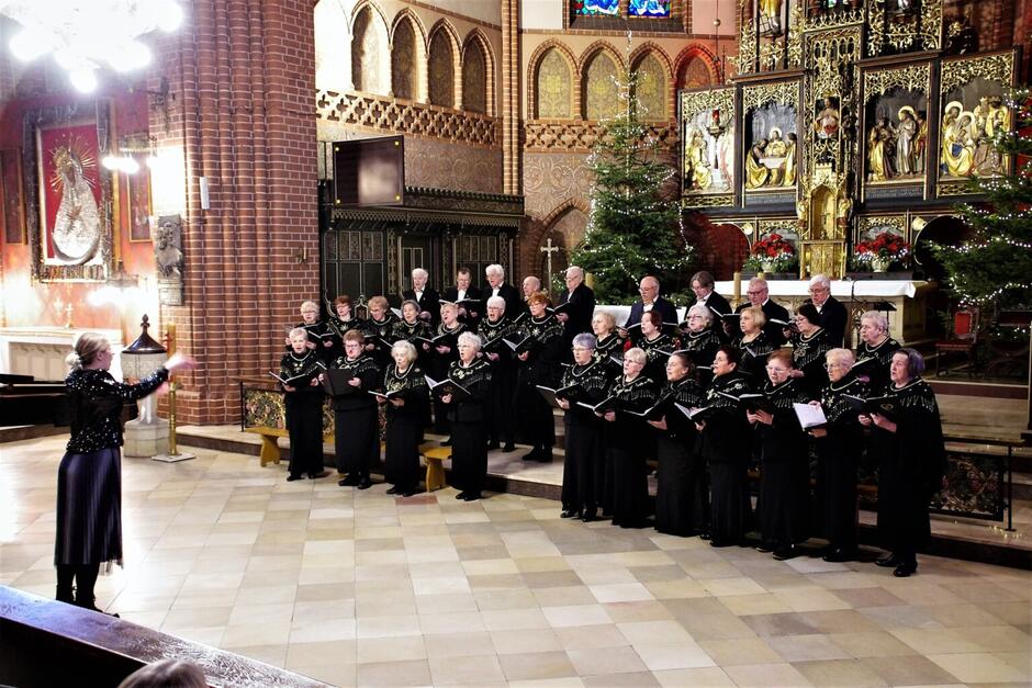 Chór występuje w kościele przed ozdobionym ołtarzem, wykonując kolędy pod batutą dyrygentki. Świąteczny nastrój podkreślają choinki i złote detale wystroju wnętrza.
