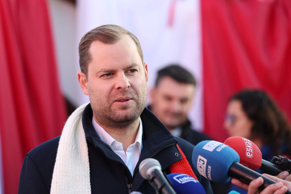 na zdjęciu mężczyzna w średnim wieku, w ciemnym płaszczu i białoczerwonym szaliku