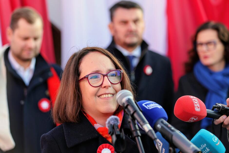 na zdjęciu kobieta w średnim wieku, w okularach, mówi do mikrofonów