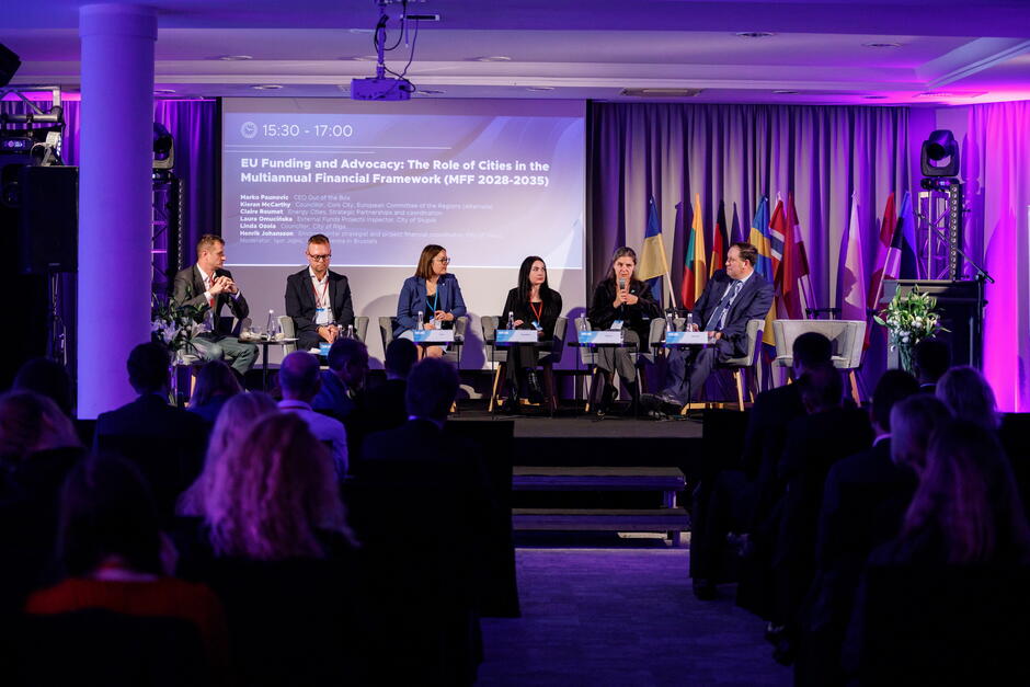 Zdjęcie przedstawia panel dyskusyjny poświęcony roli miast w nowej perspektywie finansowej Unii Europejskiej na lata 2028–2035. Na scenie siedzi sześcioro prelegentów omawiających tematy związane z finansowaniem i współpracą samorządów, a wydarzenie odbywa się w sali oświetlonej fioletowym światłem i udekorowanej flagami państw europejskich.
