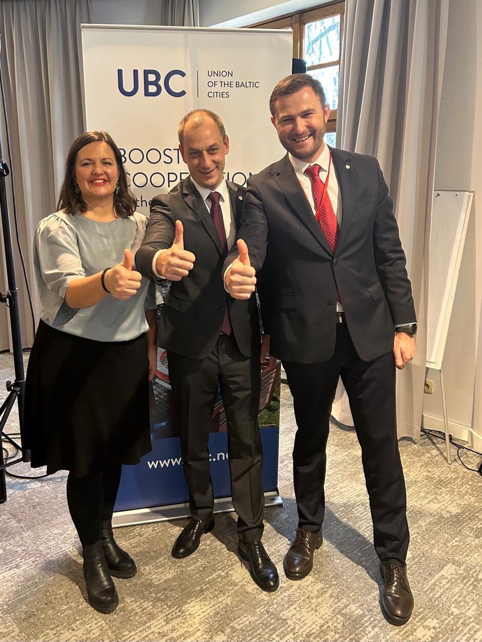 Na zdjęciu trzy osoby stoją obok siebie, uśmiechnięte i pokazujące gest uniesionych kciuków. W tle widoczny jest baner organizacji Union of the Baltic Cities, co sugeruje, że fotografia została wykonana podczas wydarzenia związanego ze współpracą miast regionu Morza Bałtyckiego.
