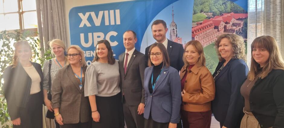 Na zdjęciu grupa uczestników pozuje wspólnie na tle banera XVIII Zgromadzenia Ogólnego Union of the Baltic Cities. Uczestnicy, ubrani formalnie, stoją uśmiechnięci w dobrze oświetlonej sali, co nadaje fotografii charakter pamiątkowy i oficjalny.
