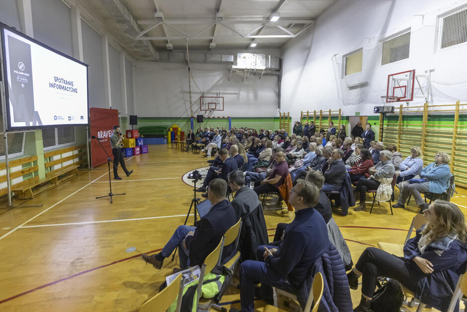 Tłum ludzi siedzi na krzesłach w sali gimnastycznej szkoły, przed nimi stoi mężczyzna i mówi do mikrofonu.