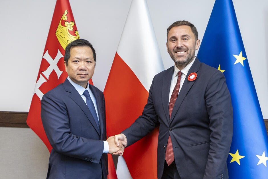 Zdjęcie dwóch mężczyzn witających się uściskiem dłoni, w tle są duże stojące flagi Gdańska, Polski i Unii Europejskiej