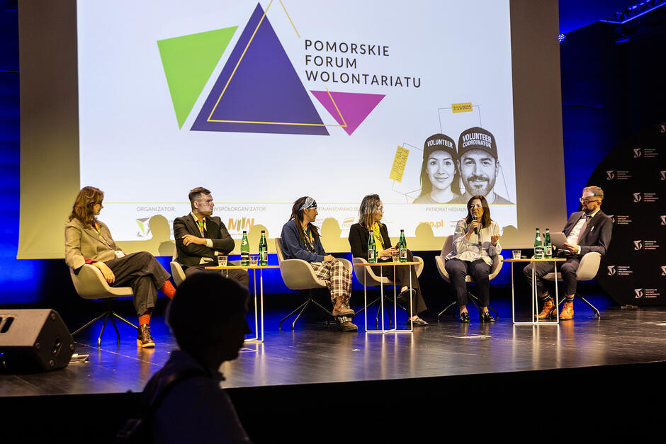 Sześć osób siedzi na fotelach przed małymi stolikami na scenie i rozmawia, nad nimi ekran z napisem Pomorskie Forum Wolontariatu .