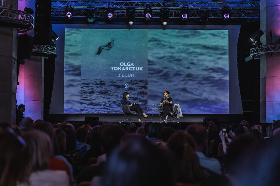 Dwie kobiety rozmawiają na scenie w gotyckim kościele, przed nim siedzi tłum słuchaczy