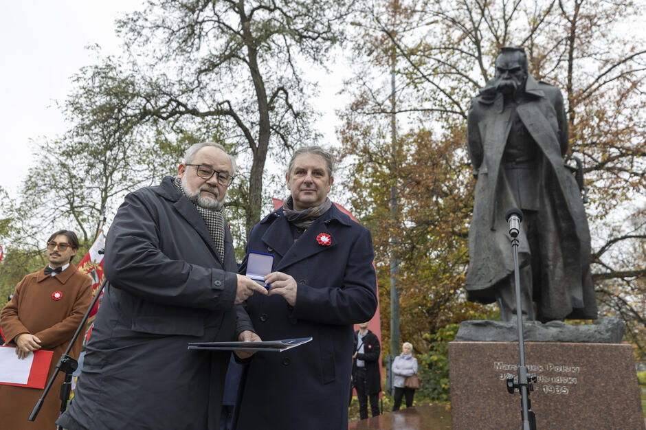 Podczas uroczystości przy pomniku Józefa Piłsudskiego odbyło się wręczenie odznaczenia jednemu z uczestników wydarzenia. W tle widoczny jest monument marszałka oraz zgromadzeni uczestnicy obchodów.

