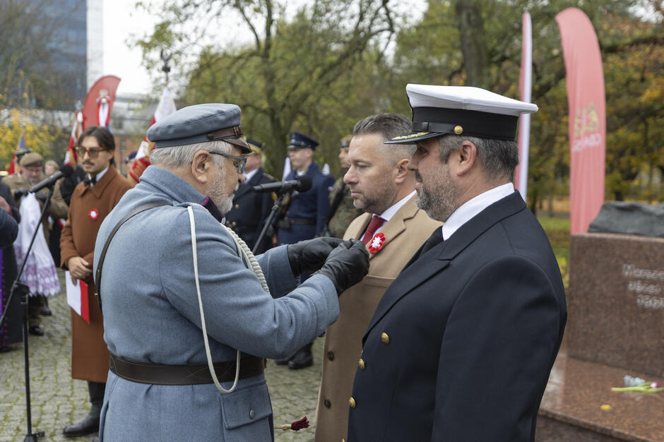 Podczas uroczystości patriotycznej przed pomnikiem Józefa Piłsudskiego jeden z uczestników w historycznym mundurze przypina odznaczenia dwóm mężczyznom. W tle widać poczty sztandarowe, przedstawicieli służb mundurowych oraz zebranych uczestników wydarzenia.
