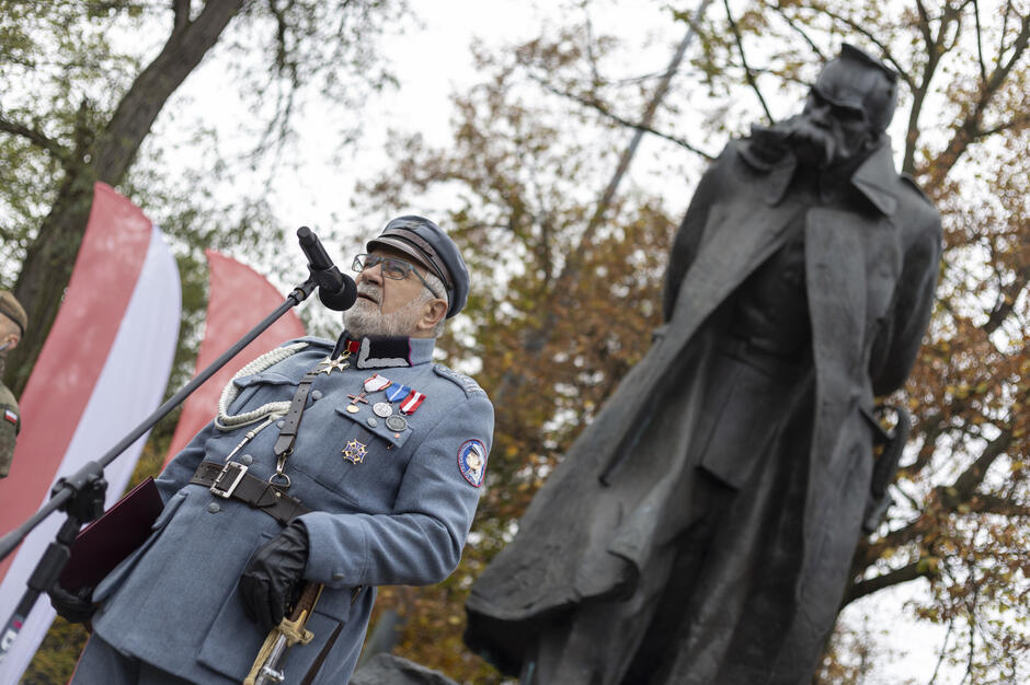 Mężczyzna w historycznym mundurze legionowym przemawia przy mikrofonie podczas uroczystości przed pomnikiem Józefa Piłsudskiego. Za nim widoczny jest monument marszałka oraz biało-czerwone flagi ustawione wśród jesiennych drzew.
